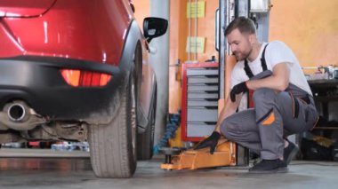 A skilled man in a uniform fixes a car, he examines it from all sides. A professional car mechanic at a service station