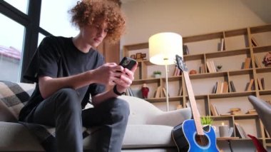 A young guy with curly hair is sitting on a sofa and holding a smartphone in his hands. Use of mobile Internet