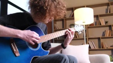 Creative young curly guy musician playing melody on acoustic guitar while sitting on sofa. 4k video