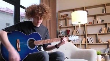 A pretty young man plays the guitar while sitting on a sofa in a bright living room.