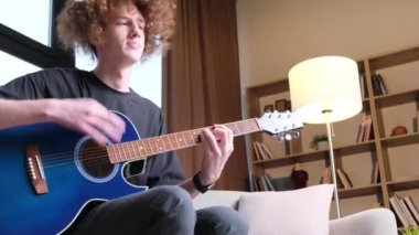 Young handsome man sitting on sofa and playing acoustic guitar in stylish room. The blogger is learning to play the guitar. 