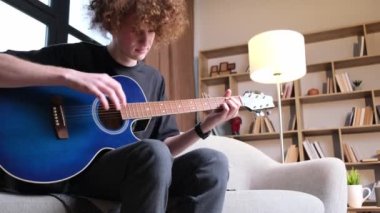 A curly-haired guy is sitting on a sofa and playing an acoustic guitar, he has a headache. Migraine and headache in a young man