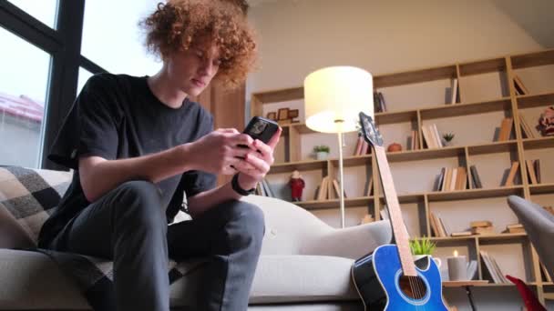 Young Guy Curly Hair Sitting Sofa Holding Smartphone His Hands — Vídeos de Stock
