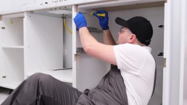 A qualified craftsman in special clothes installs the doors of a white cabinet in a modern kitchen. Installation of fittings in a new house. Home renovation concept