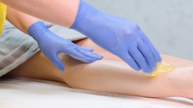The procedure for removing hair from a womans body. Hands of a female beautician in blue gloves apply cosmetic wax to a clients legs in a modern spa salon.