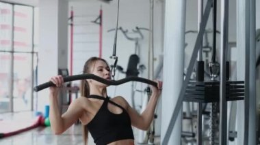 Persistent woman doing workout on the simulator. Athletic woman trains muscles by pulling a barbell on herself in the gym.