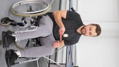 Vertical video. A young man of European appearance in a wheelchair plays with a racket and a ping-pong ball. A disabled person is preparing for a game. Paralympian