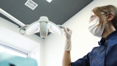 Portrait of a female dentist in a mask before starting dental treatment. The dentist adjusts the lighting lamp. Painless dental treatment