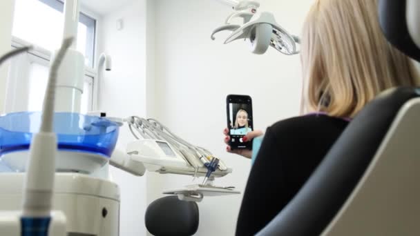 歯の椅子の若いブロンドは スマートフォンからインターネットを介して通信します 入院中の歯科治療前の女性患者 — ストック動画