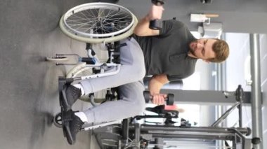 Vertical video of young attractive bearded man in wheelchair lifting dumbbells in gym. Rehabilitation after injuries, damage to the lower limbs. Life with disabilities