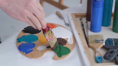The hand of a young artist mixes paints on a palette that stands on the table. Basic colors. Work with a thick paintbrush. Workplace of the artist