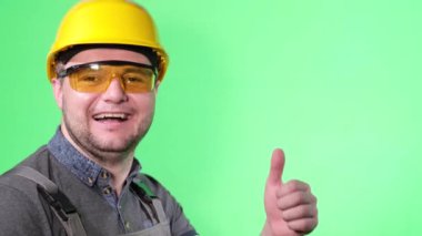 Happy smiling worker showing thumb up on green background in studio. Advertising shooting of a young builder with a European appearance.