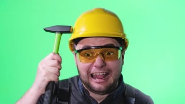 A madly angry construction worker bangs a hammer on his hard hat. Crazy angry construction worker on green background in photo studio.