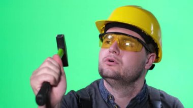 A young concentrated worker in a hard hat and glasses on a green background reproduces the knocking of a hammer. Concentrated worker with hammer.