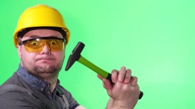 A madly surprised strained worker in a helmet and glasses on a green background reproduces the knock of a hammer. A builder with big eyes and a puffy mouth. Crazy facial expressions