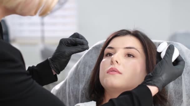 美容室で若いブルネットに永久的な眉メイクを行う手袋の女性のクローズアップ 眉の輪郭 現代の化粧品 顔の美容ケア — ストック動画