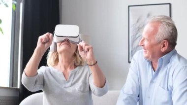 Beautiful elderly couple with gray hair having fun with virtual reality headset sitting on sofa at home. Happy grandparents use modern technology