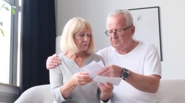 Serious elderly husband and wife review utility bills. A stressed elderly couple is sitting at the table at home, analyzing financial documents.