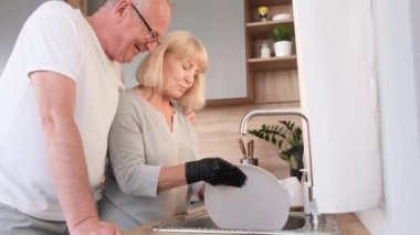 Happy elderly couple washing dishes in the kitchen after dinner, loving elderly man hugging his wife from the back.