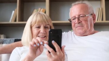 Smiling senior couple using smartphone browsing internet together sitting on sofa at home and hugging. Elderly couples use a modern gadget.