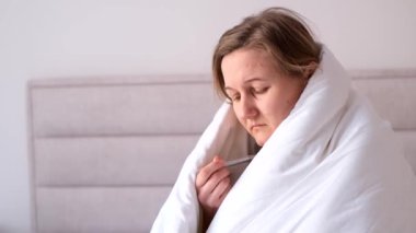 Portrait of sick depressed woman suffering from cold, measuring temperature with thermometer, sitting in bed covered with blanket at home.