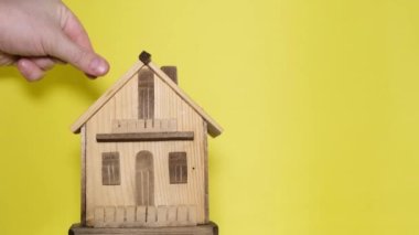 A mans hand fills a piggy bank in the shape of a house on a yellow background. Saving money on building a house concept.