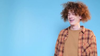 Curly red-haired student on a beautiful blue background is posing and gesturing with his hands. Advertising shooting in the studio