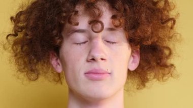Close-up of the face of a curly red-haired boy, a student on a light brown background. The emotion of peace, happiness. Relaxation, meditation. A guy with narrowed eyes.