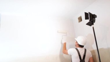A young man in a helmet and uniform paints the white walls in the apartment with a roller, using a lamp and a spotlight for lighting. Modern renovation in the apartment