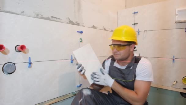 一个面带微笑的男人穿着特殊制服 戴着保护眼镜 在浴室里修补瓷砖的画像 补偿者 砖头安装程序 — 图库视频影像