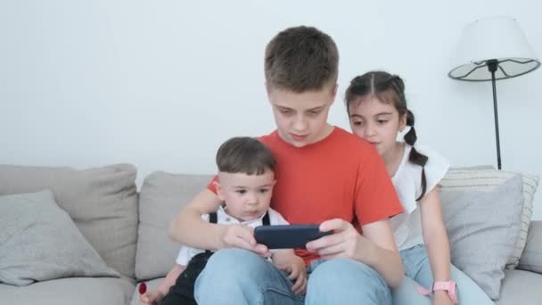 Τρία Παιδιά Διαφορετικών Ηλικιών Χρησιμοποιούν Smartphone Ενώ Κάθονται Στον Καναπέ — Αρχείο Βίντεο