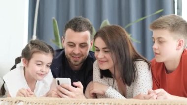 A cheerful family lying on the sofa together. The father is smiling and sharing a moment with his two kids using a smartphone. They are happy and using the modern gadget together
