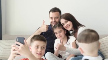 Mutlu bir çift ve üç çocuğu kanepede oturuyor. Ebeveynler birlikte gülümser ve akıllı telefondan fotoğraf çekerler. Ailedeki neşeli ebeveynler ve çocuklar
