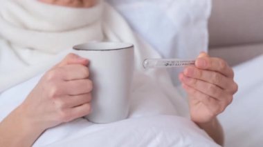 Hasta kadının elinde bir fincan beyaz çay tutarken ve termometreyle sıcaklığı ölçerken yakın plan çekimi. Evde SARS tedavisi.