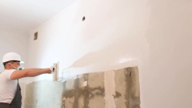 A repairman in a mask and helmet paints the wall white. Modern renovation in the apartment