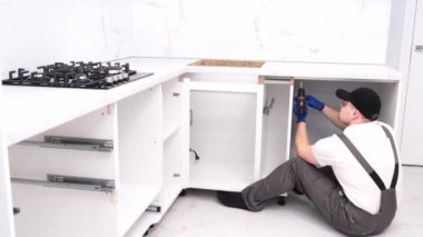 A craftsman in overalls installs kitchen furniture and accessories while sitting on the floor. The worker uses tools
