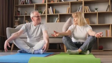 Oturma odasındaki spor paspaslarının üzerinde yoga egzersizleri yapan yaşlı ve güzel bir çift. Emeklilikte sağlıklı bir yaşam tarzı