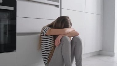 Mutfak odasında yerde oturan yorgun genç bir kadın, başı ağrıyor ve üzgün, üzgün, mutsuz hissediyor..
