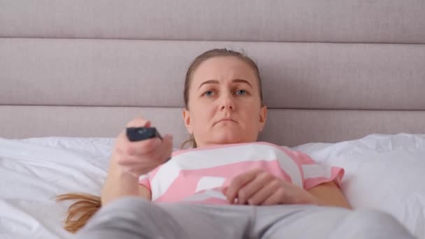 リモコンを操作しながらテレビを見ている疲れた女性 彼女は頭痛がして動揺してベッドに行く — ストック動画