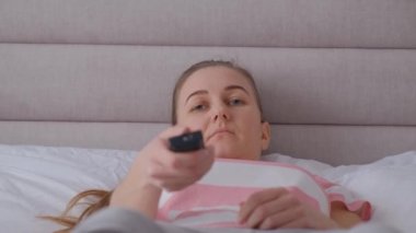 Avrupalı görünümlü genç bir kadın elinde uzaktan kumandayla yatakta uzanıyor ve sıkıcı bir şekilde kanalları değiştiriyor. Depresyondaki kadın TV ile yatakta vakit geçiriyor..