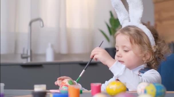 イースターのためにウサギの耳をした美しい小さな女の子 うさぎの耳をした笑顔の女の子がイースターのために卵をペイント 幸せな子供 — ストック動画
