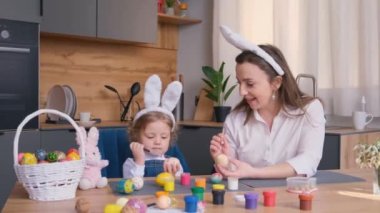 Genç bir anne ve küçük sevimli tavşan kulaklı kızı Paskalya yumurtalarını bir sepet ve bir tavşanla bir şenlik masasında otururken boyuyorlar..