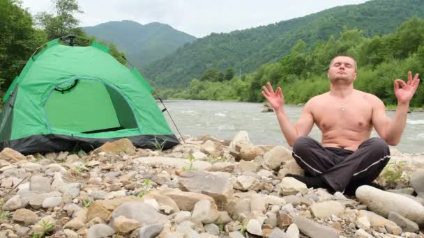 若い男は 山脈の美しい景色の真ん中に苦い川のほとりに座っている間 ヨガを練習します 美しい場所で休んでください 捨てられた場所 — ストック動画