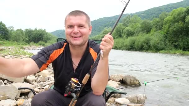 山の川の近くに座って釣竿とハンサムな内容の若い漁師の肖像画 美しい山の風景の中で釣り — ストック動画