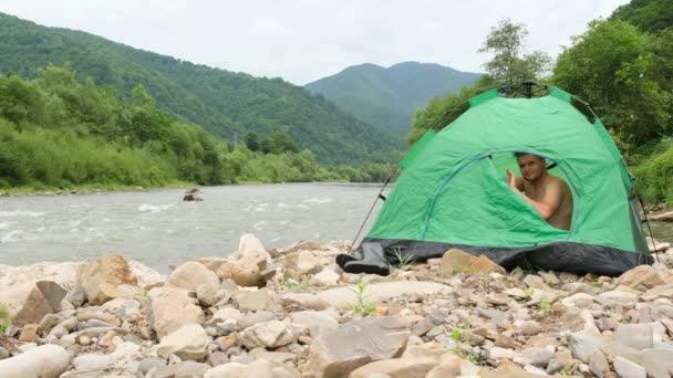 一个年轻人在山河岸边打开一个绿色的帐篷 挥挥手 满意的游客 — 图库视频影像