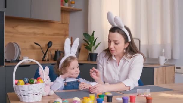 イースターの家族の伝統 若い母親を愛することは 台所のテーブルに座っている間 イースターのための塗料で卵をペイントし 飾るために小さな子供を教えています — ストック動画
