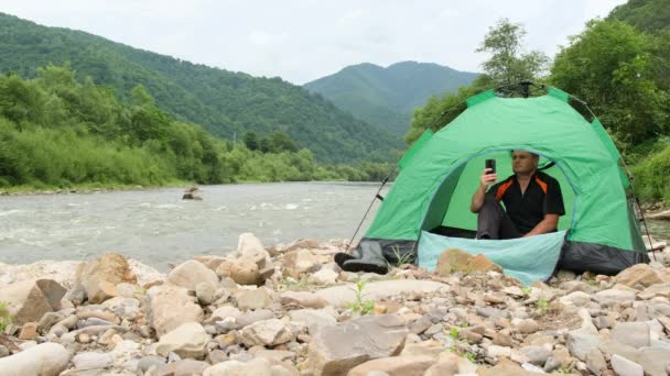 若い観光客が川の土手のテントに座り 彼の携帯電話で写真を撮る 自然の中でのレクリエーションの概念 — ストック動画