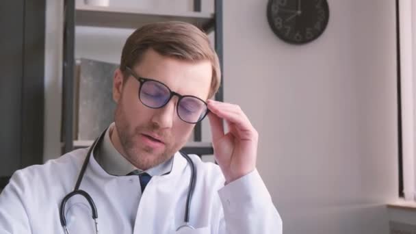 留着胡子 身穿白衣的医生摘下眼镜 坐在办公室的桌子旁 工作中的问题 诊所的疲劳 — 图库视频影像