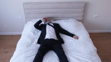 Siyah takım elbiseli yorgun, rahat bir adam büyük beyaz bir yatakta uzanıyor ve telefonda konuşuyor. Yorucu bir iş gününden sonra rahatlama