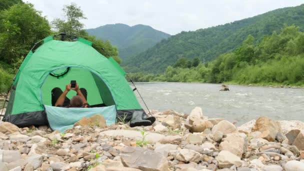 観光客は 電話を見て 山の川の銀行の緑のテントの中にあります 山の真ん中の新鮮な空気の中で休む — ストック動画